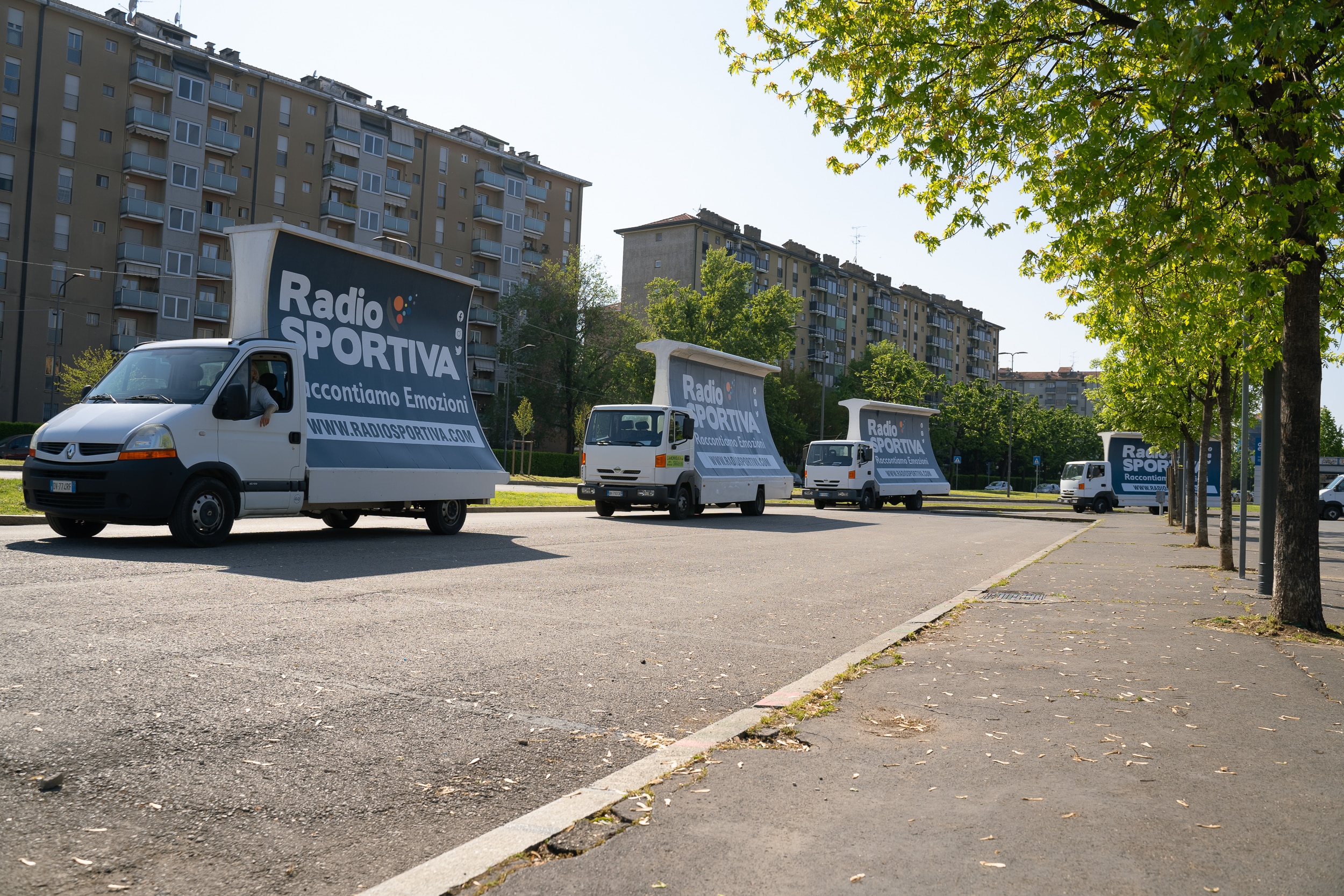 Fila di camion vela Radio Sportiva parcheggiati lungo un viale alberato